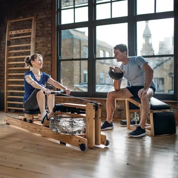 WaterRower Nohr rowing machine with light wood frame and clear tank, alongside a man with dumbbells in a minimalist modern interior.