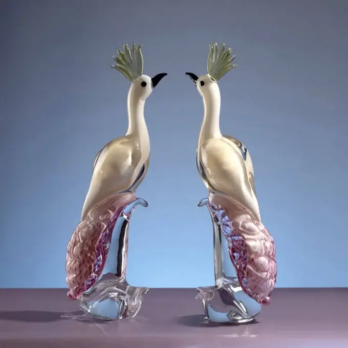 Crystal peacock sculptures, clear glass with pearlescent white bodies, pink & purple filaments, on mauve surface against blue-lavender gradient.