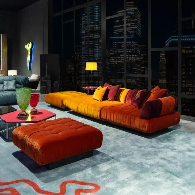 Burnt orange velvet modular sofa with deep cushions, paired with a matching ottoman on a blue rug, alongside a blue sofa and neon sculpture.