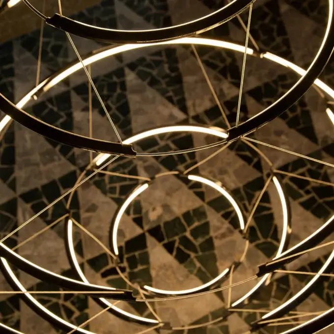 Concentric ring chandelier, dark metal finish, layered illumination above dark gray and beige mosaic tile surface.