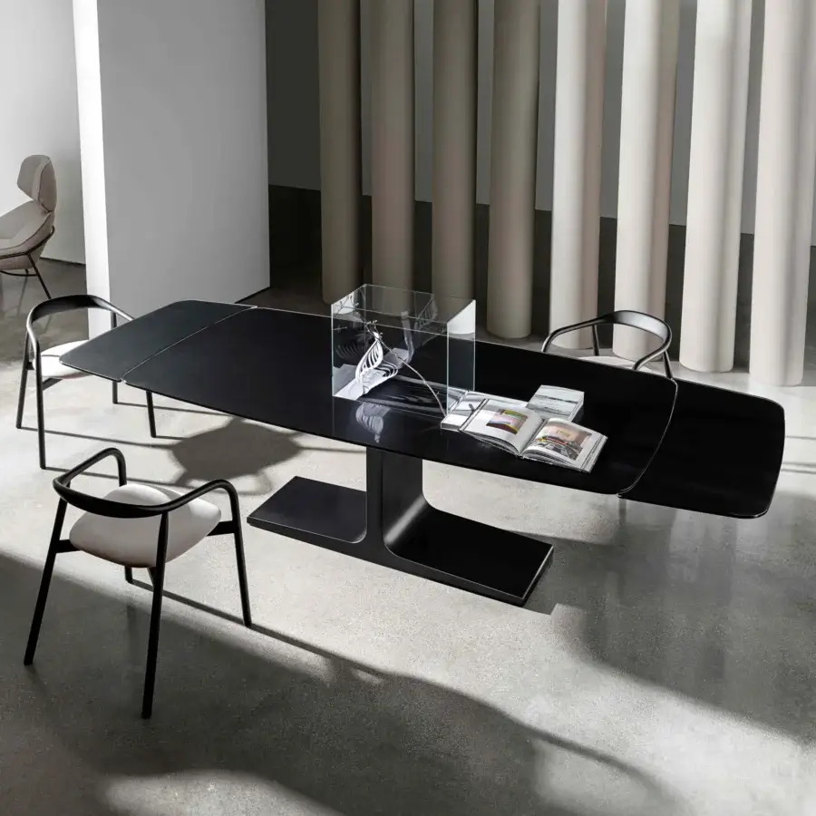 Black extendable table with glass top and dark gray fabric panels, supported by curved metal base, in modern interior setting.