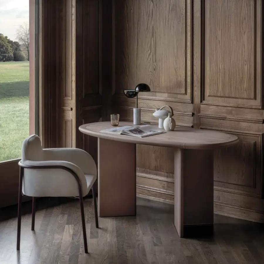 Zed desk, taupe finish, oval tabletop, with dark burgundy chair, bronze and ceramic lamps, in room with wood paneling.