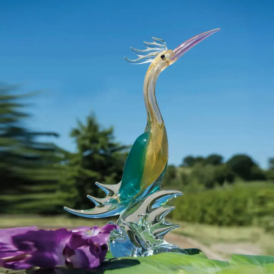 Glass paradise bird sculpture, teal to gold, with slender neck and wave-shaped base, displayed with foliage against blurred trees.