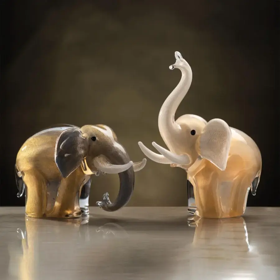 Glass elephant sculptures, amber-gold and creamy beige, facing each other with curved trunks on a light-grey reflective surface.
