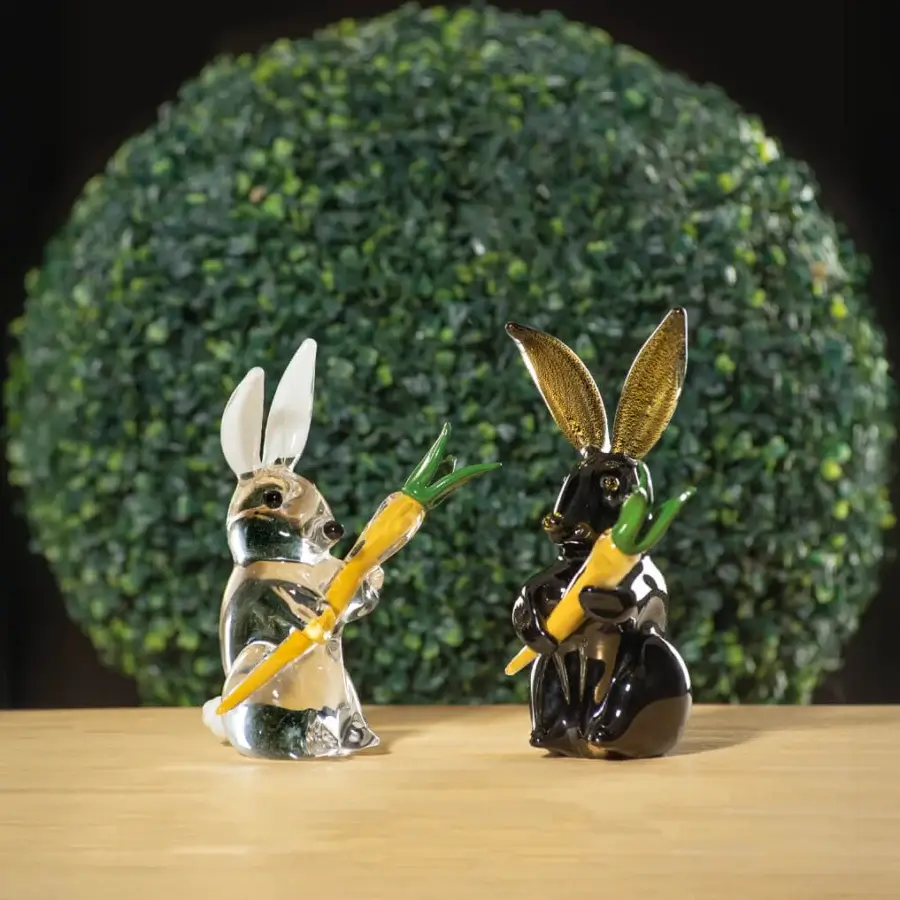 Glass rabbit sculptures, transparent & black, with gold ears, holding carrots, displayed on wood before a green foliage wall.