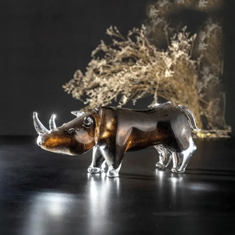 Glass rhino sculpture, amber-toned, walking stance on black reflective surface with golden dried foliage backdrop.