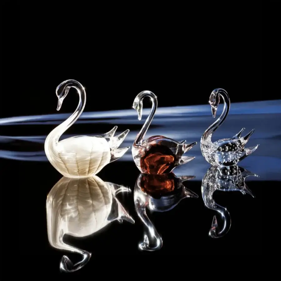 Glass swan sculptures by Duccio Di Segna—white, amber, and clear—with curved necks displayed on a reflective black surface, showcasing glass textures.