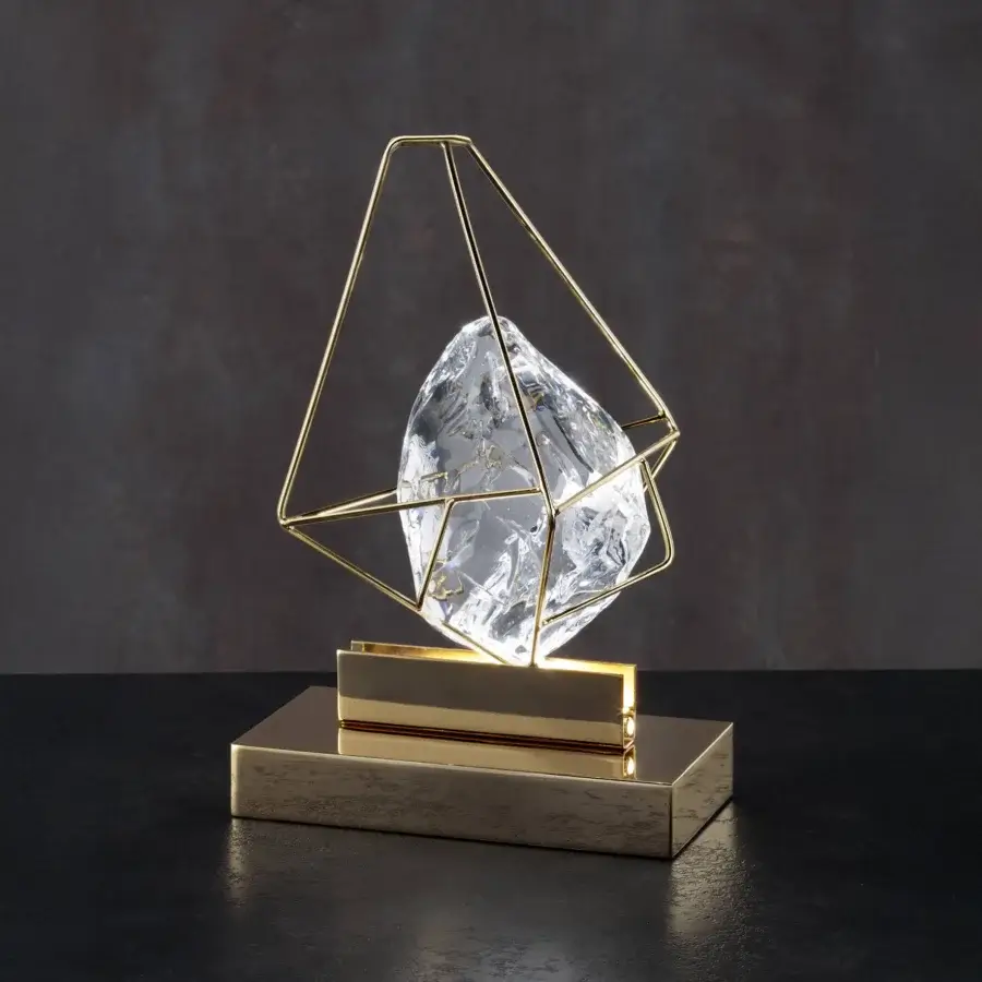 Crystal table lamp, brushed gold metal cage, truncated pyramid shape, clear faceted crystal, dark gray background, reflective black surface.
