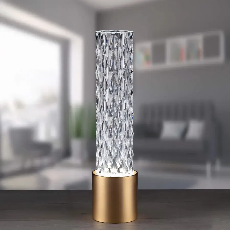 Cassoni table lamp, clear faceted shade, brushed brass base, illuminated against blurred gray interior with sofa and geometric art.