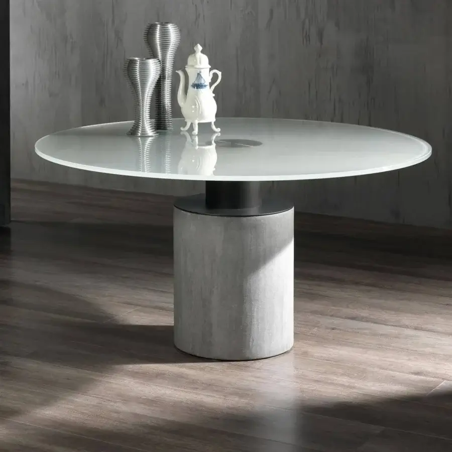 Translucent circular surface on a two-part cylindrical pedestal displays reflected shapes and a white ornate vessel against a gray backdrop