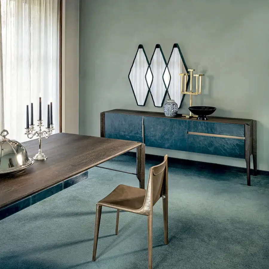 Teal sideboard with brass trim, paired with dark wood dining table, light brown leather chair, geometric metal wall art, turquoise carpet.