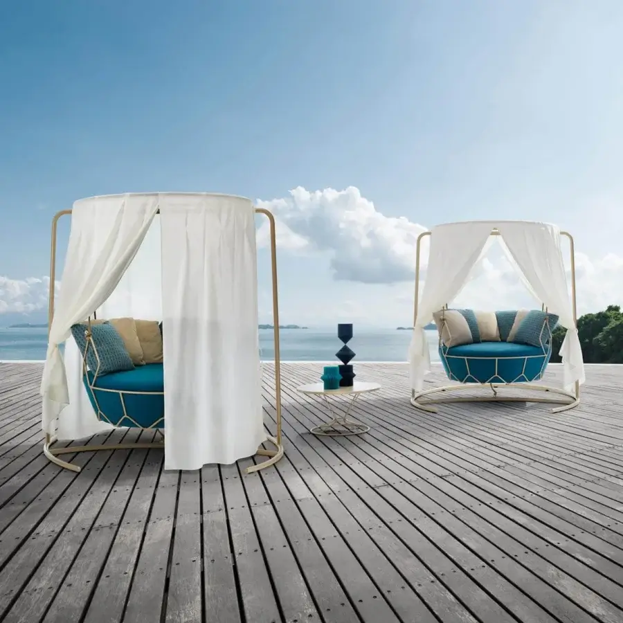 Lavish Roberti Rattan Gravity swing lounge chair with teal cushions, a sheer white sunshade and a light golden powder-coated steel structure