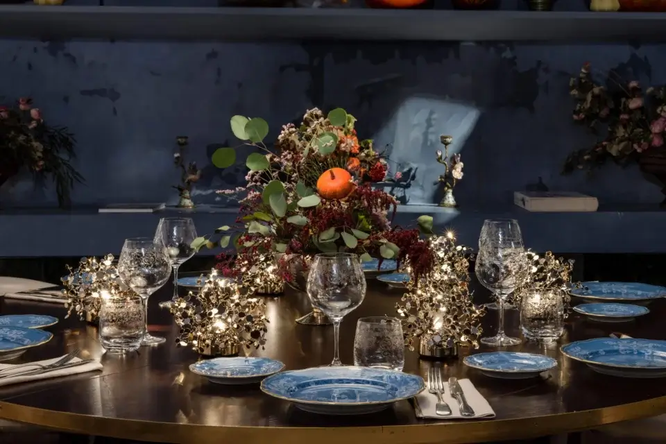 Round dining table with dark wood surface and bronze base set for Thanksgiving, featuring blue patterned plates, floral centerpiece, and lit pumpkins above.