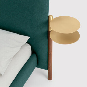 Brass tray with two circular tiers and walnut stem attached to a dark green upholstered bed frame, studio shot.