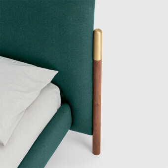 Teal upholstered headboard with reddish-brown wood and brushed brass cylindrical support, white linen pillow, light gray backdrop.
