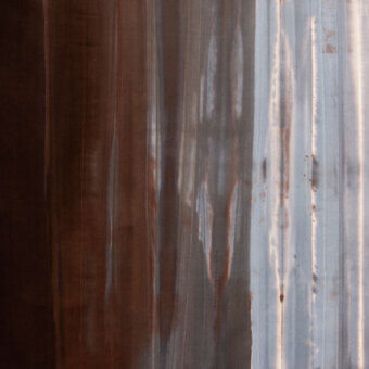 Corrugated metal surface displaying a gradient from dark brown to silvery-gray, with vertical ridges and oxidation patterns creating textural depth.