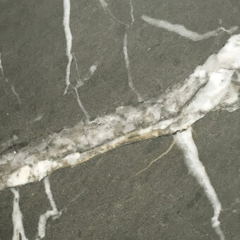 Matt Valentine Grey marble close-up with dark grey base, irregular white veining, and a diagonal fissure filled with crystalline white material.